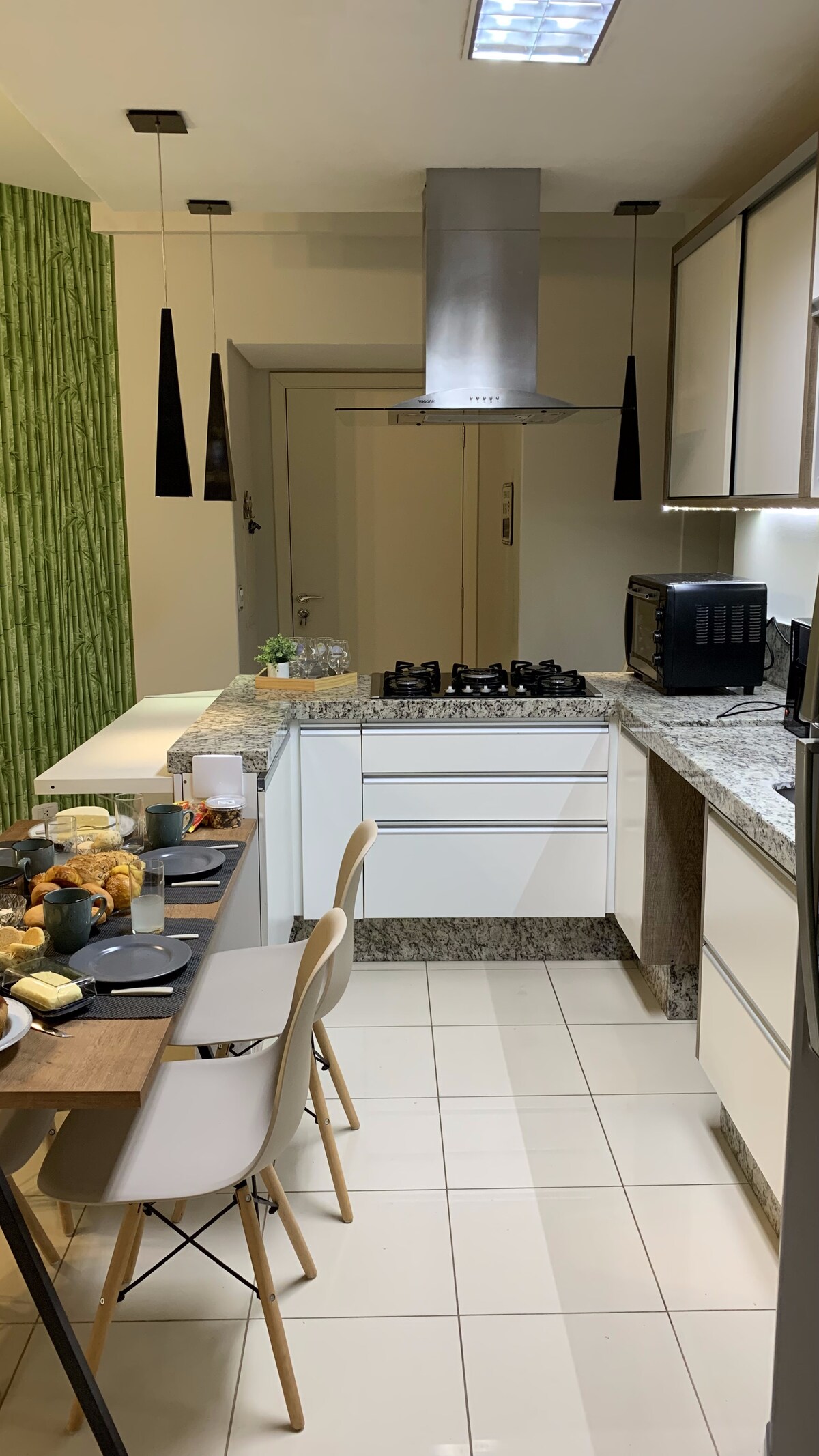A modern kitchen area features sleek white cabinetry and a granite countertop. A dining table is set with dishes and a variety of baked goods. Black pendant lights hang above the kitchen island, and a stovetop is positioned beneath a stainless steel hood.