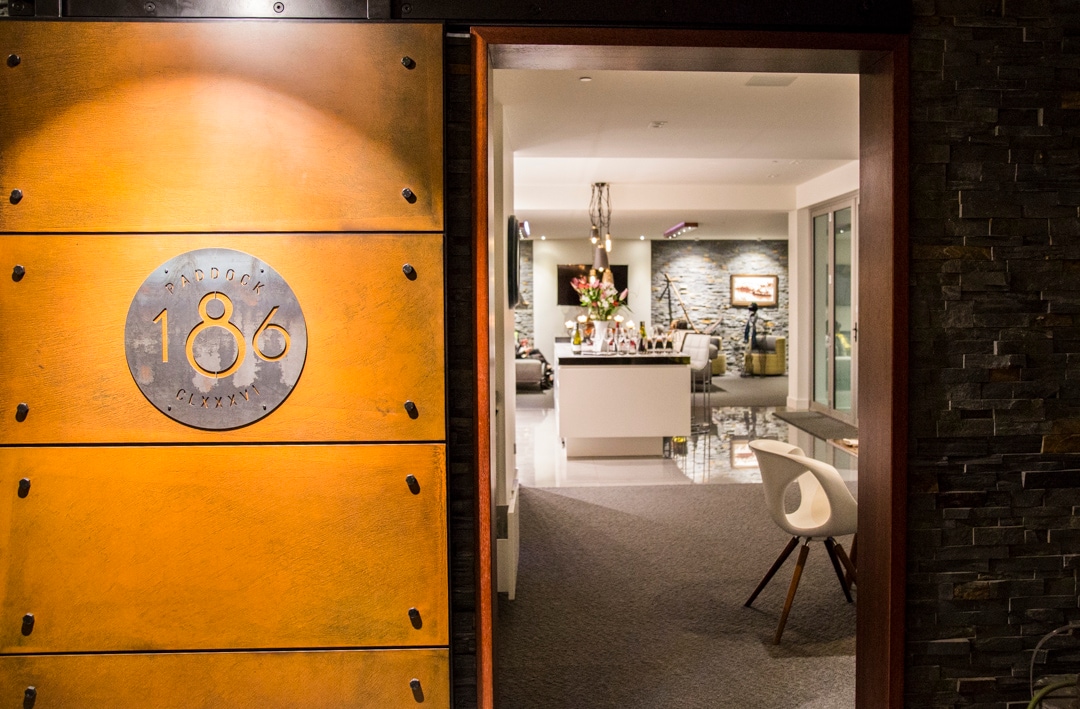 The entryway features a large, distinctive plaque with the number '186,' leading into a well-lit open space. A modern living area is visible in the background, showcasing sleek furnishings and decorative elements. The warm tones of the entrance create an inviting transition.