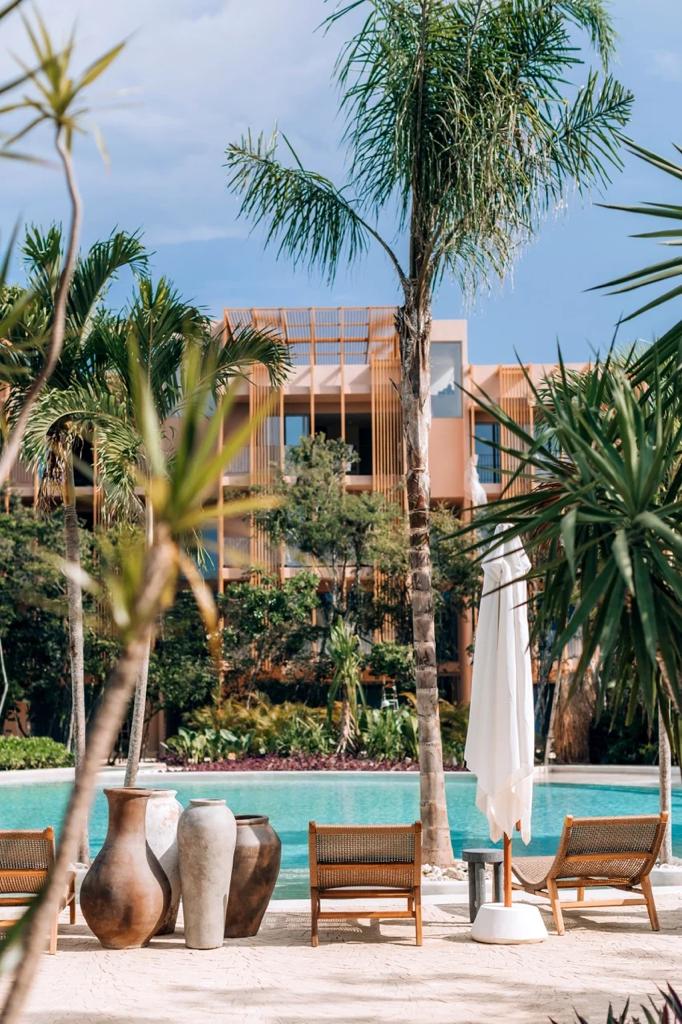 A serene pool area is framed by lush tropical plants and palm trees. Comfortable lounge chairs are placed nearby, while tall vases create an artistic touch. The inviting water reflects the soft hues of the surrounding environment, providing a tranquil backdrop for relaxation.