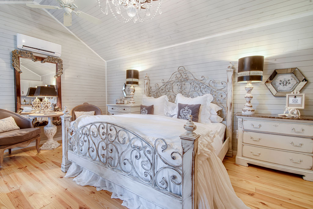 A spacious bedroom features a decorative wrought-iron bed adorned with luxury linens. Elegant nightstands are positioned on either side, complemented by stylish table lamps. A vintage mirror and armchair add character, while natural light illuminates the light-colored wooden flooring and softly textured walls.