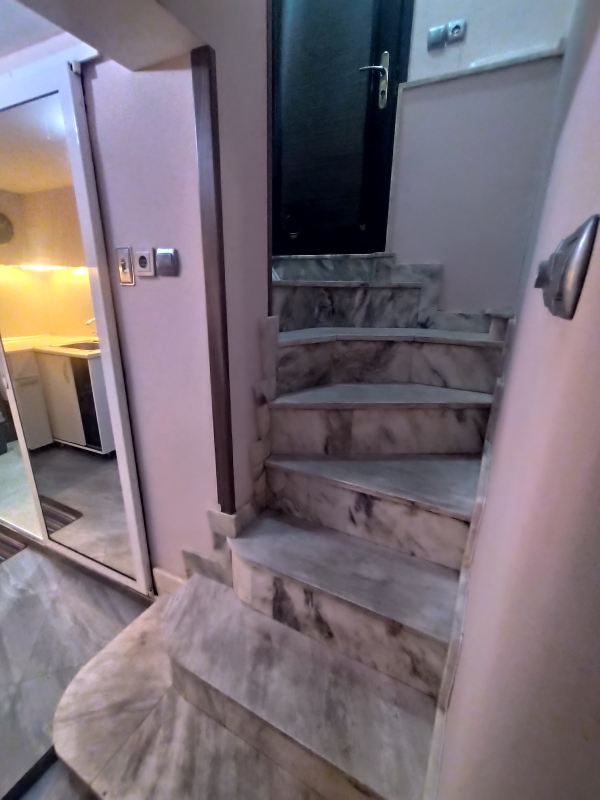 A stairway with smooth marble steps leads upwards, illuminated by soft lighting. The walls are painted in neutral tones, and a dark door is visible at the top. The kitchen area is seen in the background through a partially open doorway.
