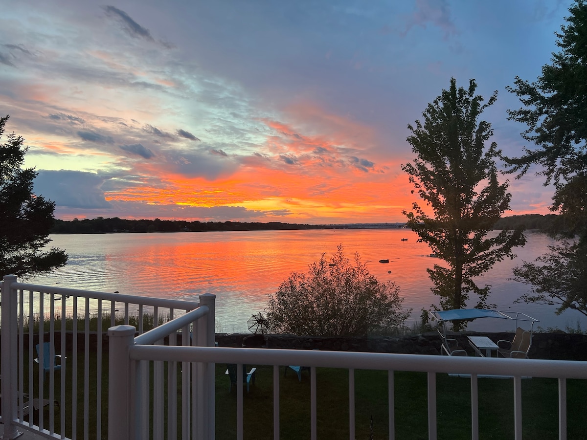 A stunning sunset is captured over the river, with vibrant colors reflecting on the water's surface. Silhouettes of nearby trees and chairs are outlined against the sky, creating a serene atmosphere. The scene is framed by a balcony railing, enhancing the tranquil view.