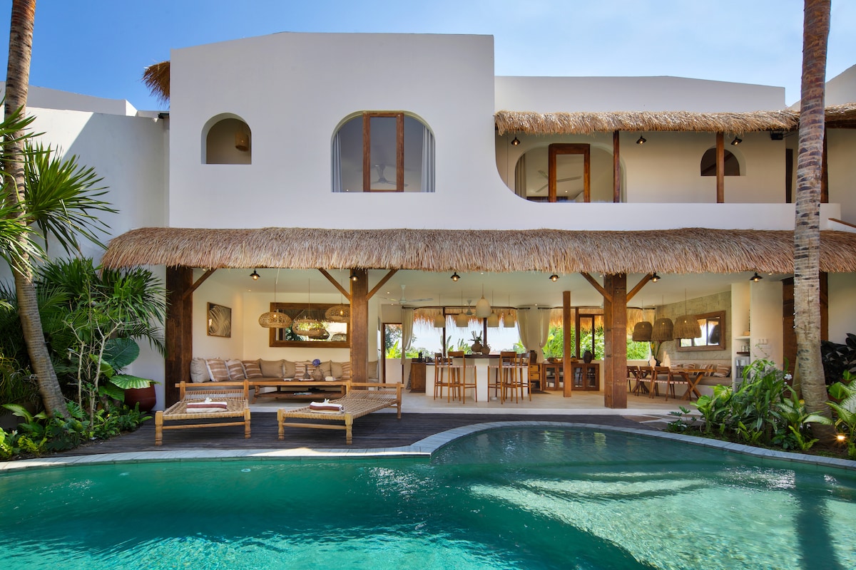 A spacious outdoor area features a private pool with clear water, surrounded by tropical foliage. The villa's architecture blends traditional Mediterranean style with Balinese elements. A thatched roof covers the terrace, where wooden lounge chairs and a dining area are visible, inviting relaxation.