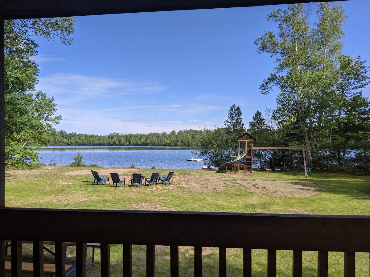 A serene view of Lake Clair is presented, featuring a grassy area with several lounge chairs arranged in a circle. In the background, a children's play area is visible alongside the calm waters, framed by trees and blue sky.