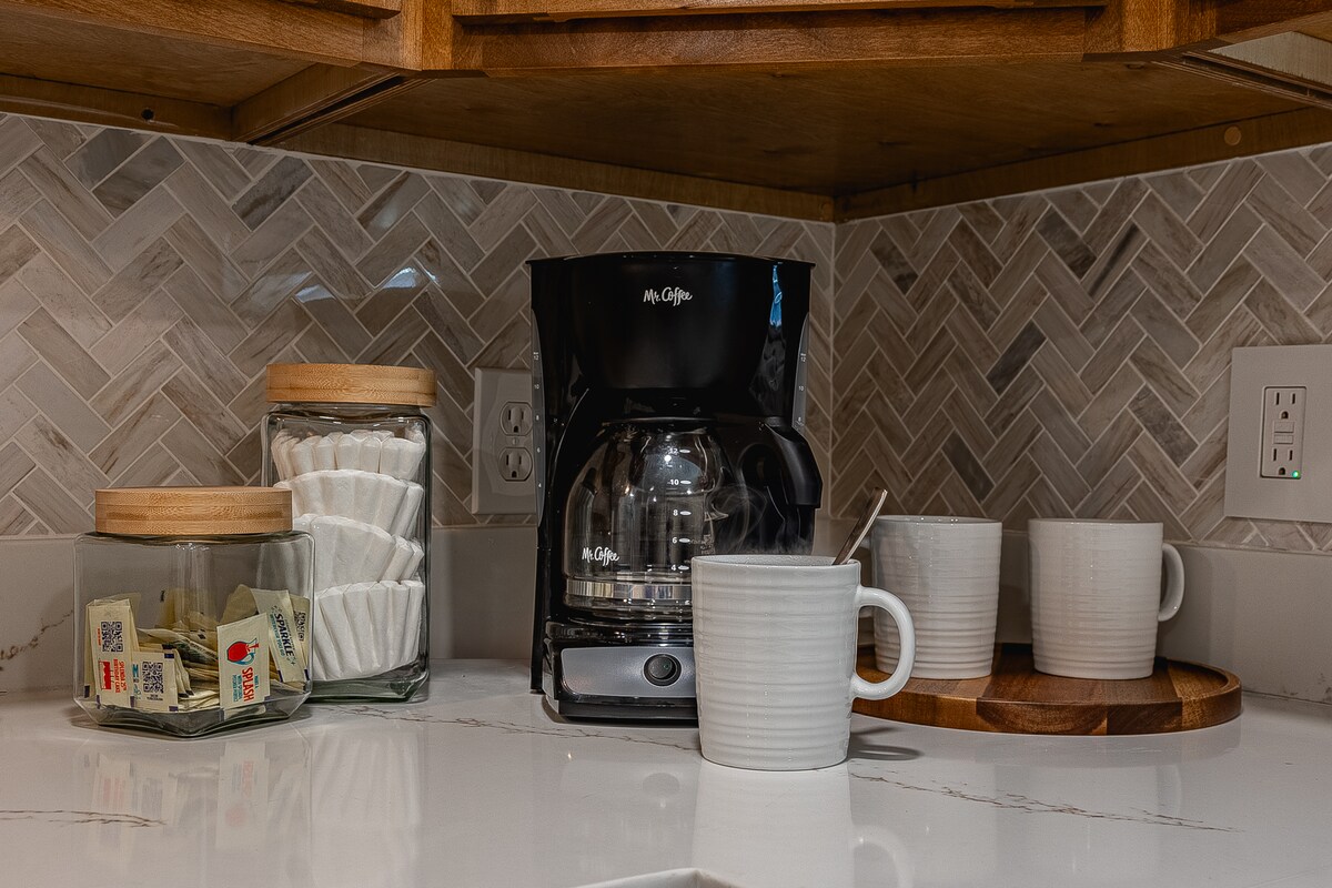 A coffee station is neatly arranged on a countertop, featuring a coffee maker and stackable mugs. Glass jars with wooden lids contain coffee filters and assorted tea bags. The backsplash showcases a herringbone tile pattern, adding a subtle texture to the setting.
