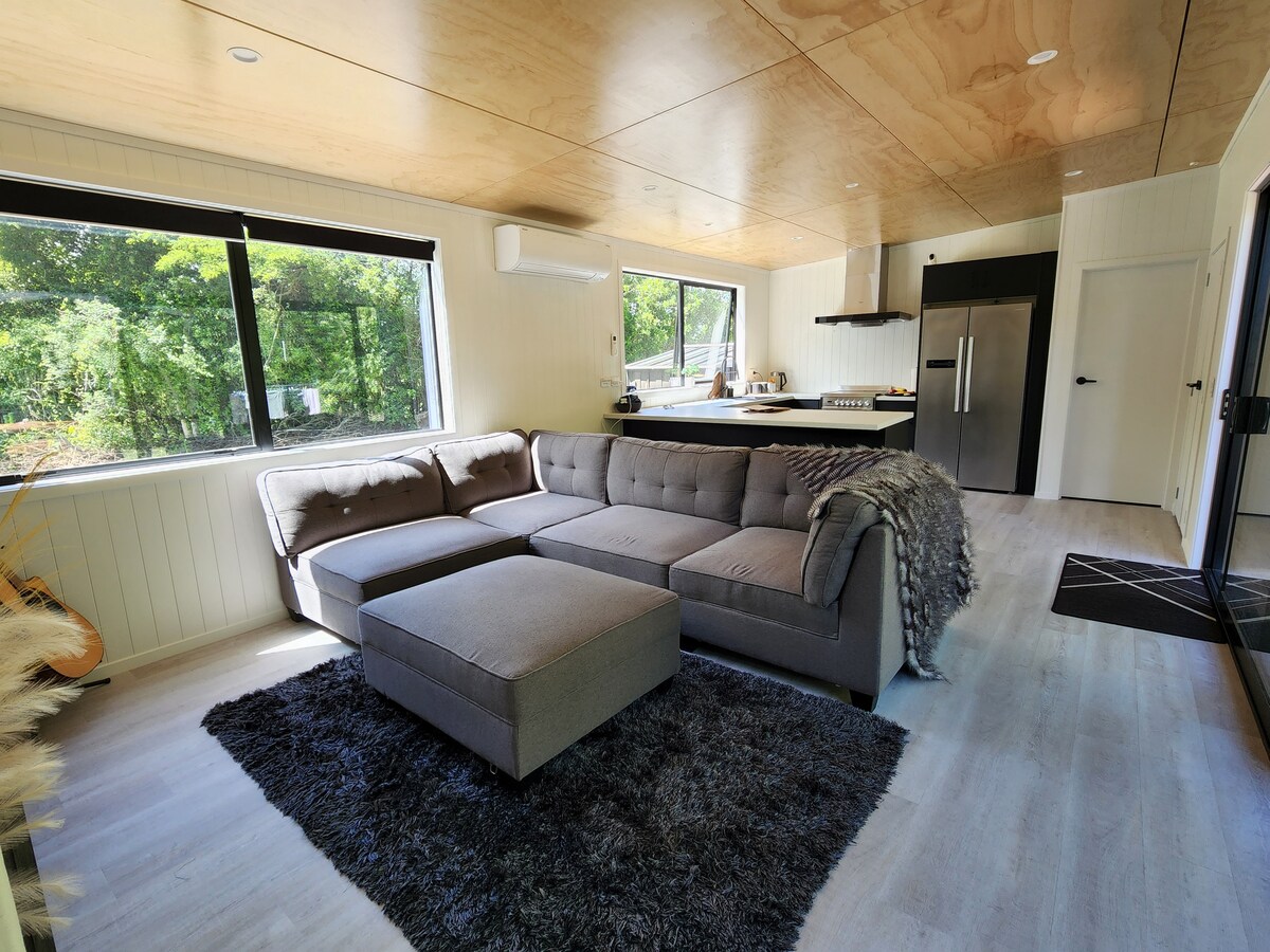 A modern living area features a gray sectional sofa positioned on a soft area rug. Large windows provide views of surrounding greenery. A sleek kitchen is visible in the background with stainless steel appliances, complemented by light-colored cabinetry and an airy atmosphere.