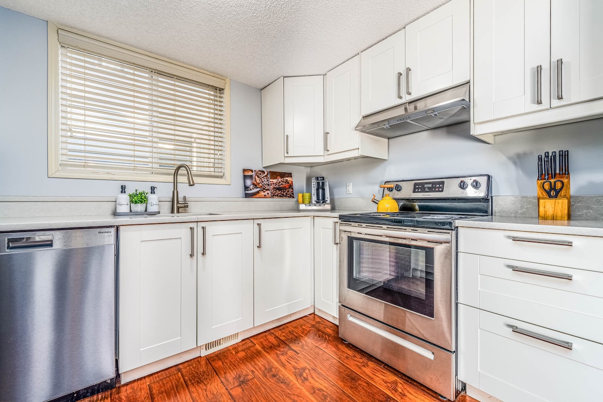 A modern kitchen is shown with sleek white cabinetry and stainless steel appliances. A window allows natural light to brighten the space, while essential cooking utensils are neatly arranged. Countertops offer ample workspace, complemented by a dishwasher and an organized stove area.