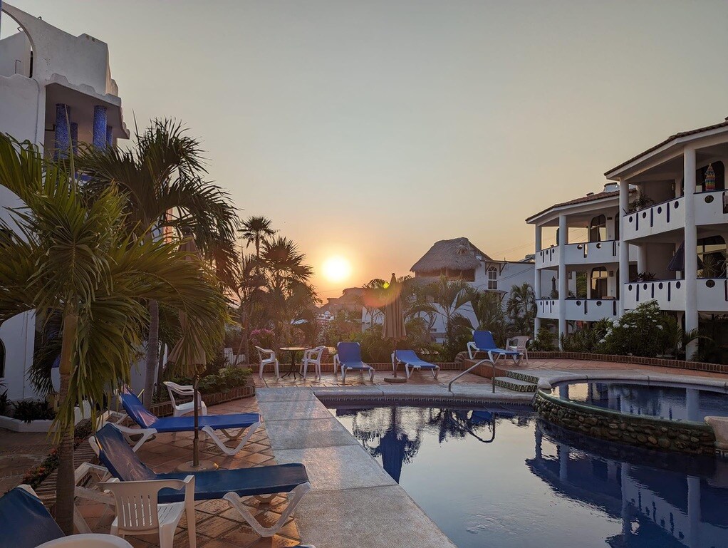 A tranquil scene captures a swimming pool surrounded by lounge chairs, with the sun setting in the background. Lush tropical plants and beautifully maintained buildings frame the area, showcasing a peaceful ambiance within the condo complex.