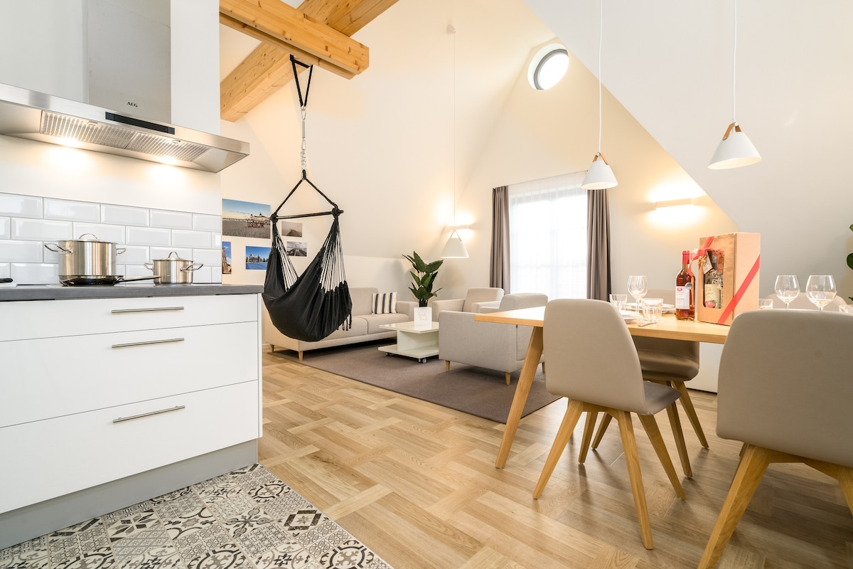 An open living space is highlighted, featuring a modern kitchen with stainless steel appliances. A cozy dining area accommodates four people, while a hammock hangs attractively in the corner. Soft lighting enhances the atmosphere, complementing the inviting seating area with a light-colored sofa.