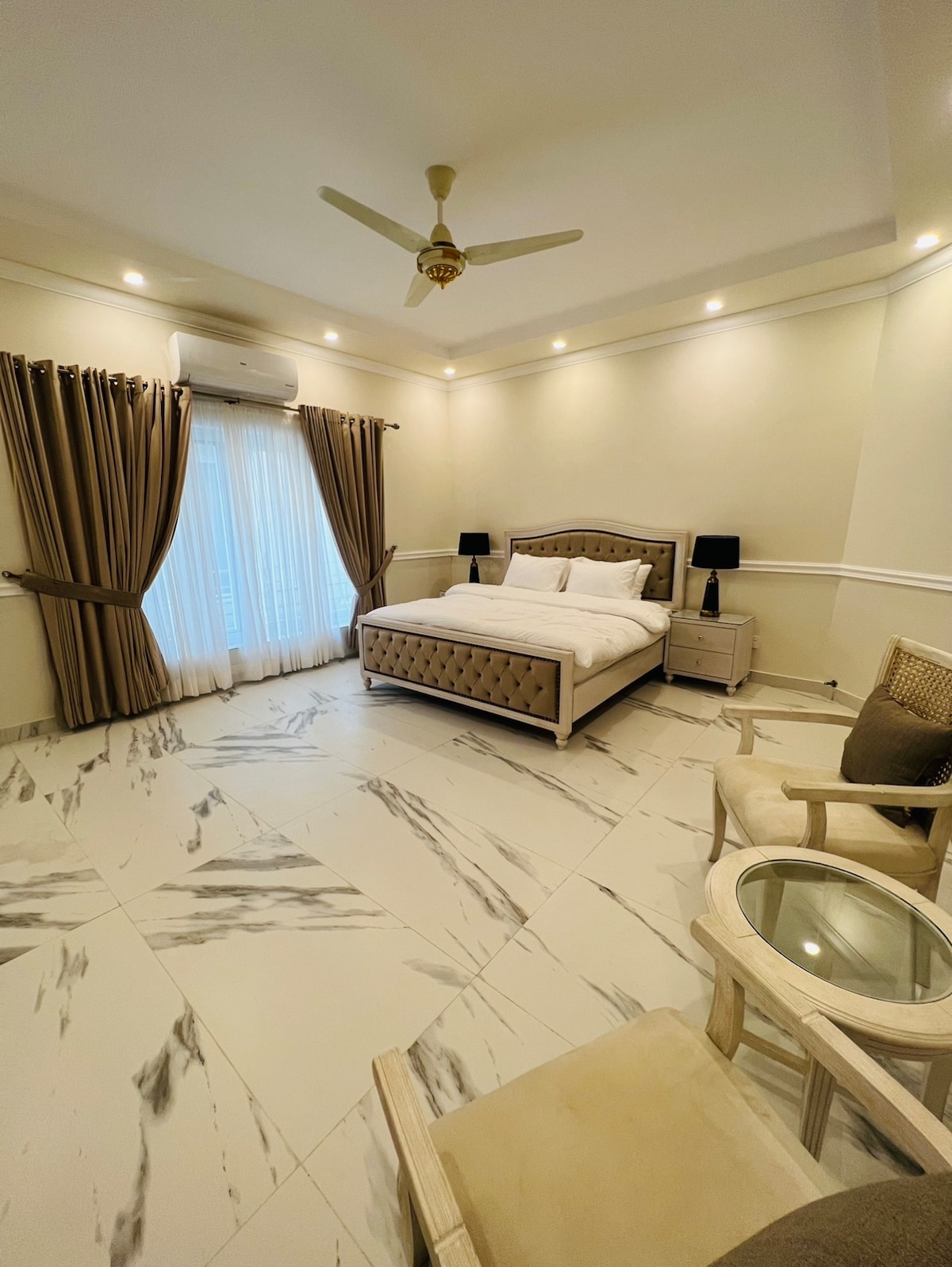 A well-appointed bedroom features a spacious bed with a tufted headboard. Natural light enters through sheer curtains. A small side table and two armchairs are positioned nearby, complemented by neutral wall tones and elegant lighting fixtures.