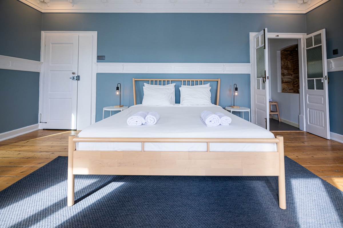 A spacious room features a light wooden bed centered against blue walls, complemented by white linens and neatly arranged towels. The room is brightened by natural light from open doors leading to a cozy bathroom. A textured area rug adds warmth to the wooden floor.