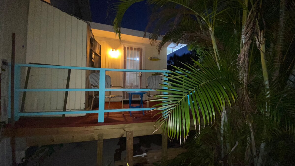 A deck area is seen surrounded by lush tropical plants, featuring two chairs and a small table. Soft lighting gently illuminates the space, complementing the serene setting in the evening. Sliding glass doors provide access to the interior, emphasizing the blend of indoor and outdoor living.