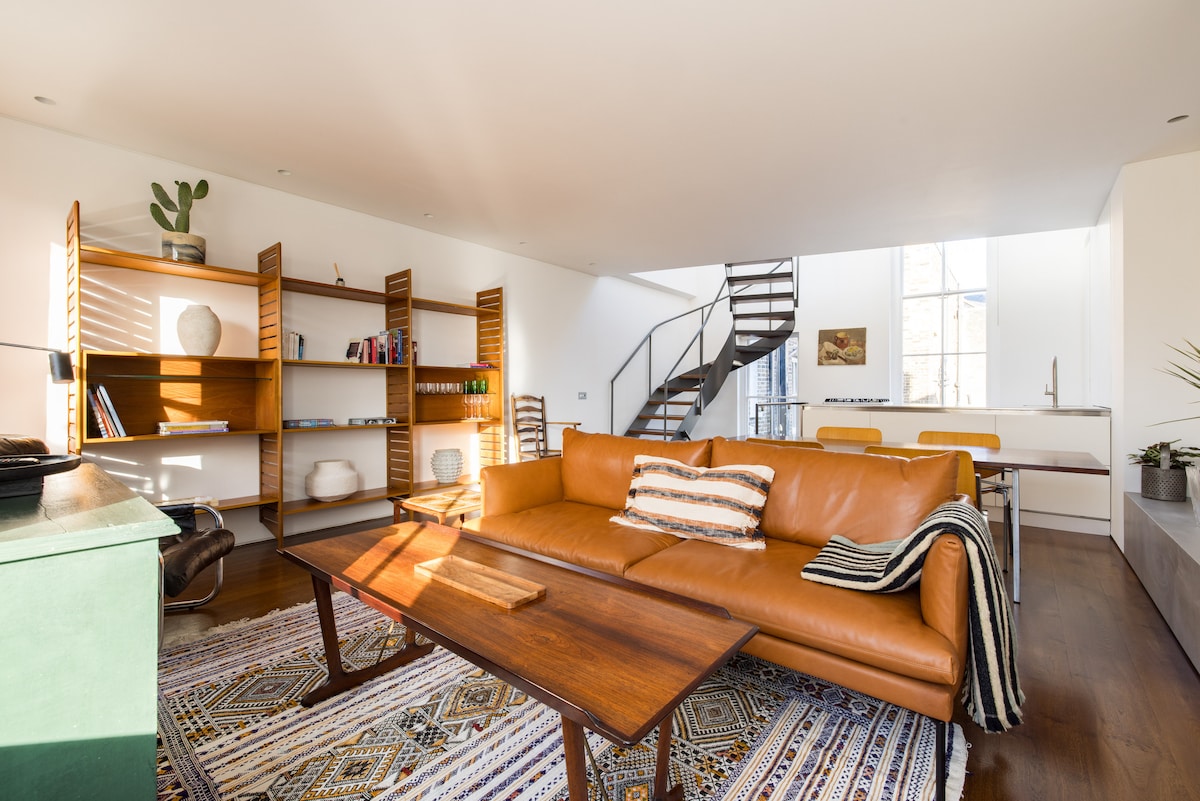 A spacious living area is filled with natural light, featuring a warm leather sofa accompanied by a patterned area rug. A wooden coffee table sits in front, while a stylish bookshelf and a staircase are visible in the background, leading to the upper level.