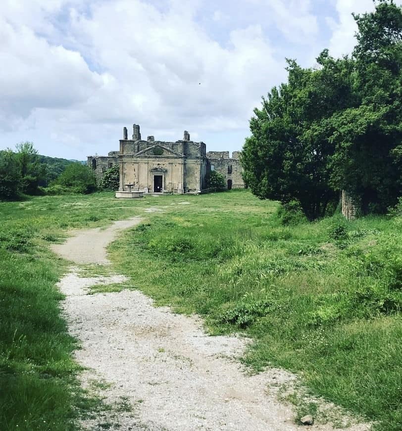 A stone building with historical architecture is visible in the distance, surrounded by lush greenery. A gravel path leads toward the structure, which features a grand entrance and remains of intricate details. Clouds drift overhead, casting soft shadows on the scene.