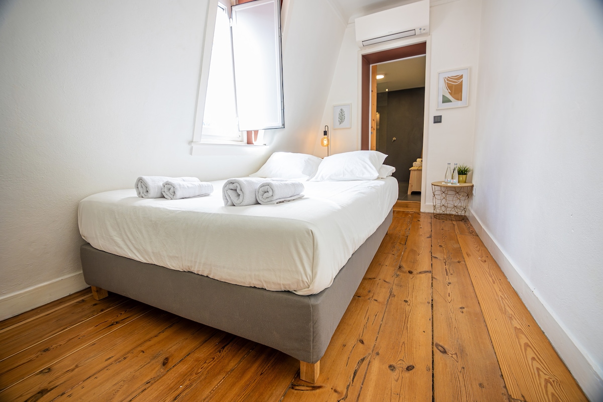 A bright bedroom features a spacious bed dressed in crisp white linens. Neatly rolled towels are placed on the bed. Light wooden flooring complements the airy atmosphere, while a window with partially open shutters allows natural light to filter in.