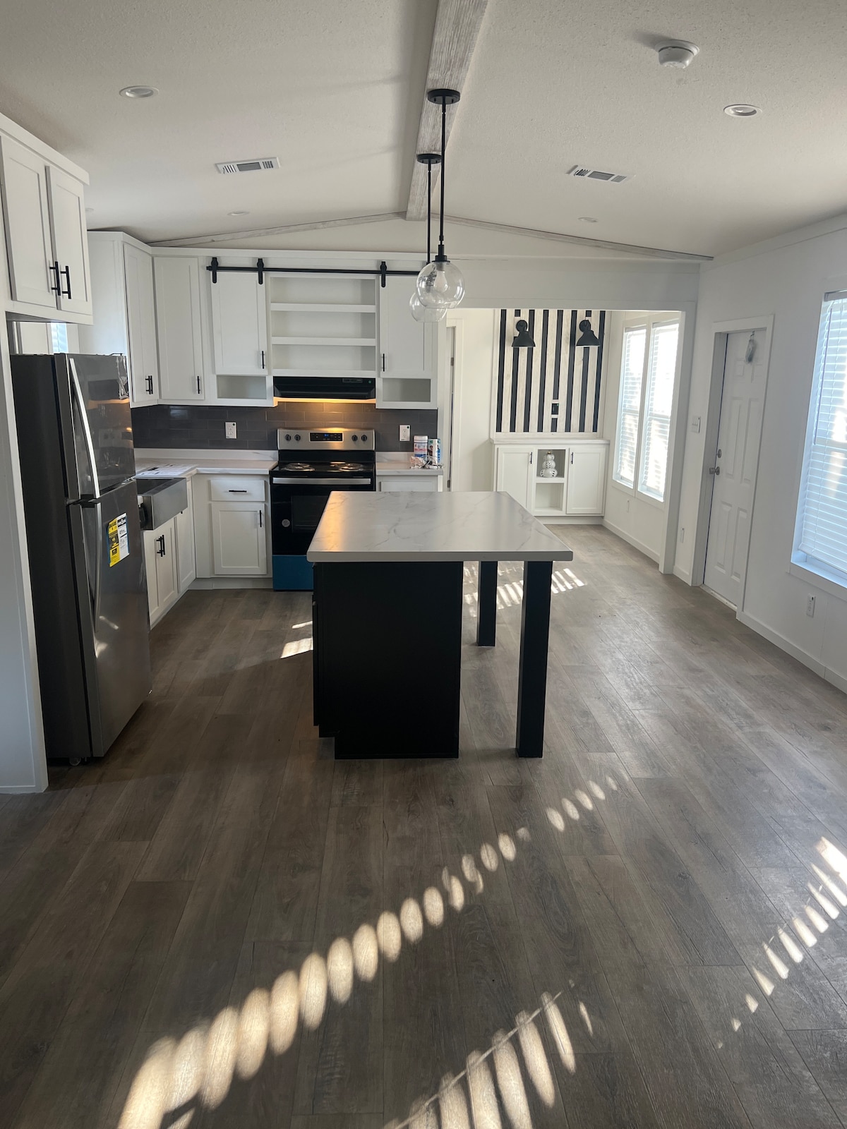 A modern kitchen is presented with sleek cabinets and stainless steel appliances. A central island with a dark finish offers additional preparation space. Bright light filters through the windows, casting patterned shadows on the floor, enhancing the open layout of the area.