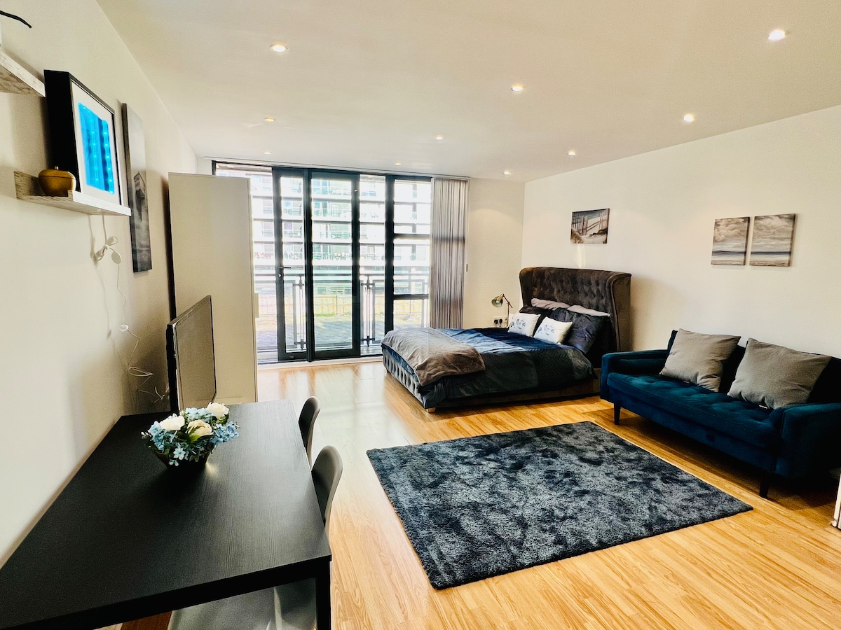 A spacious studio apartment features a comfortable sleeping area with a dark upholstered bed. A blue sofa adds to the seating options, while a sleek black table provides a workspace. Large glass doors lead to a private balcony, allowing natural light to fill the room.