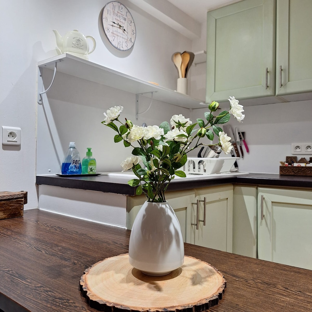 A minimalistic kitchen area features a wooden countertop with a round wooden serving board. A white vase holds fresh white flowers, adding a touch of nature to the space. Wall-mounted shelves display kitchen items, complemented by a wall clock for a functional touch.