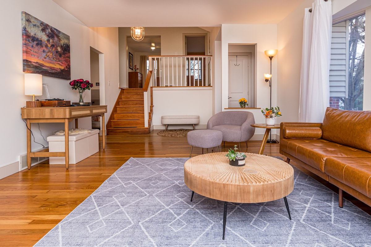 A bright living area features polished hardwood floors and a modern design. A tan leather sofa and gray armchair are positioned near a circular wood coffee table. Natural light flows in through large windows, complemented by a decorative rug and artistic wall decor.