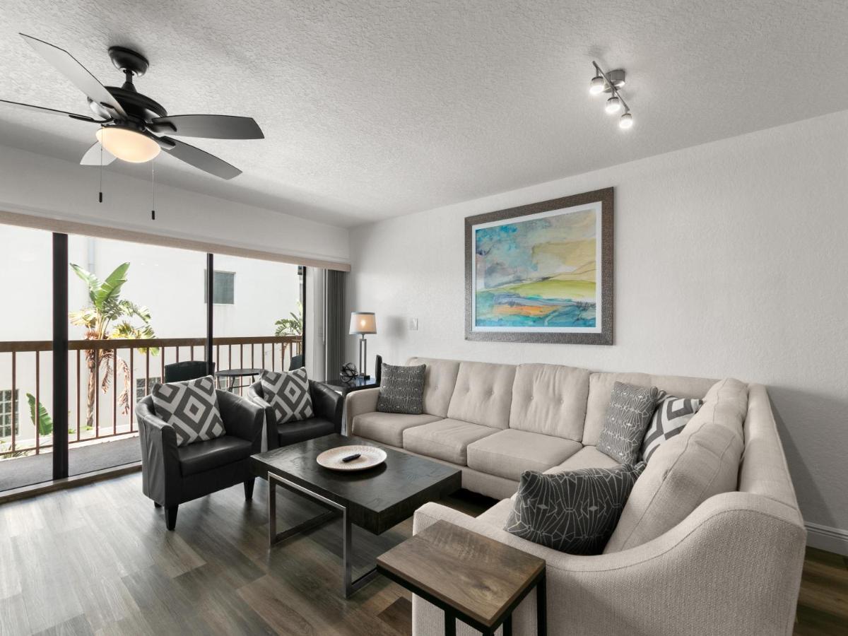 A spacious living area features a large sectional sofa with decorative pillows, paired with a coffee table. Large sliding glass doors allow natural light to fill the room, leading to a balcony with views of greenery. A ceiling fan adds a functional touch.