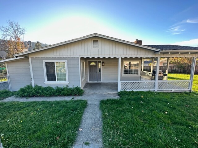 A welcoming single-story home is depicted with a well-maintained front yard featuring green grass and shrubs. The exterior includes white trim around the windows and a covered porch area. The entrance is framed by a simple, inviting door under a gabled roof.