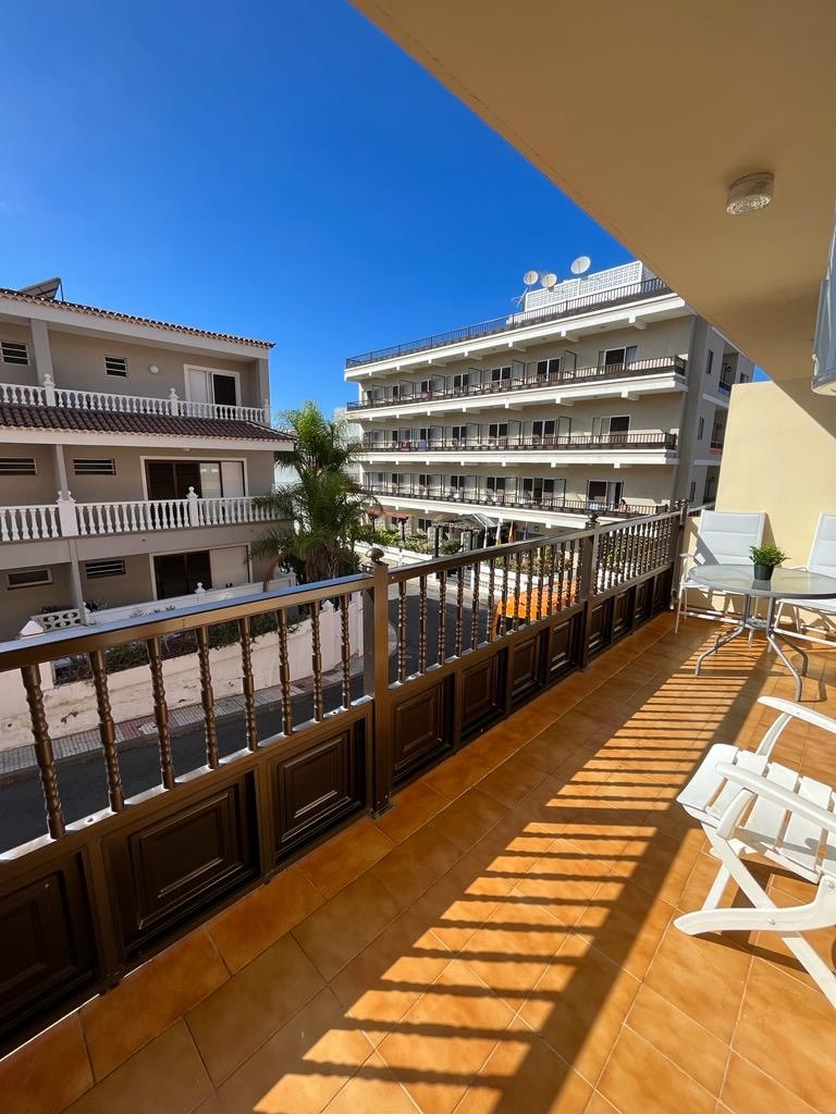 A sunny balcony offers views of surrounding buildings and palm trees. Two white loungers are positioned on the tiled floor, providing a space for relaxation. The railing features decorative wooden elements, and sunlight casts distinct shadows across the area.