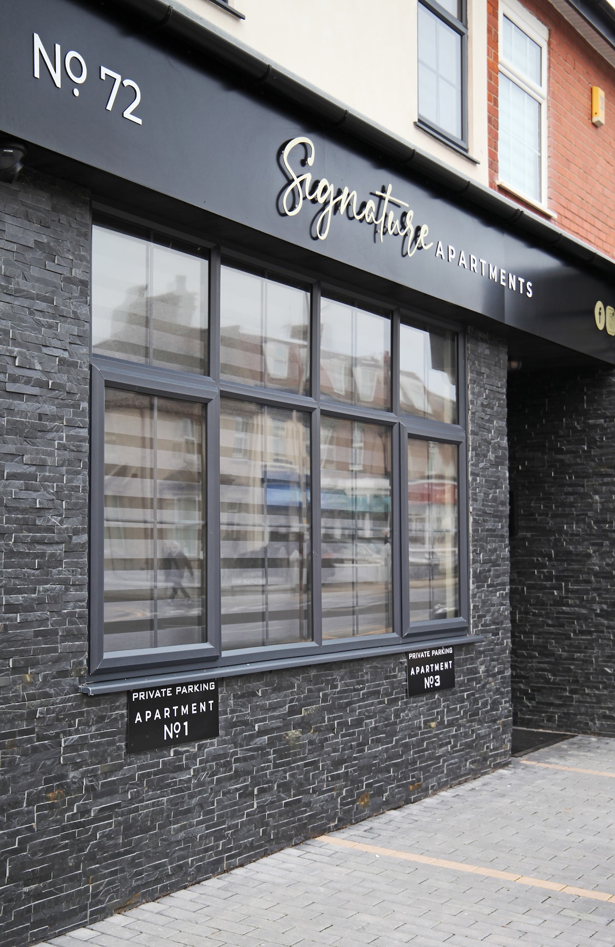 The exterior of a modern building features large windows framed in dark materials. The name 'Signature Apartments' is displayed above the entrance. Two signs indicate 'Apartment No. 1' and 'Apartment No. 3', and a paved area shows private parking nearby.