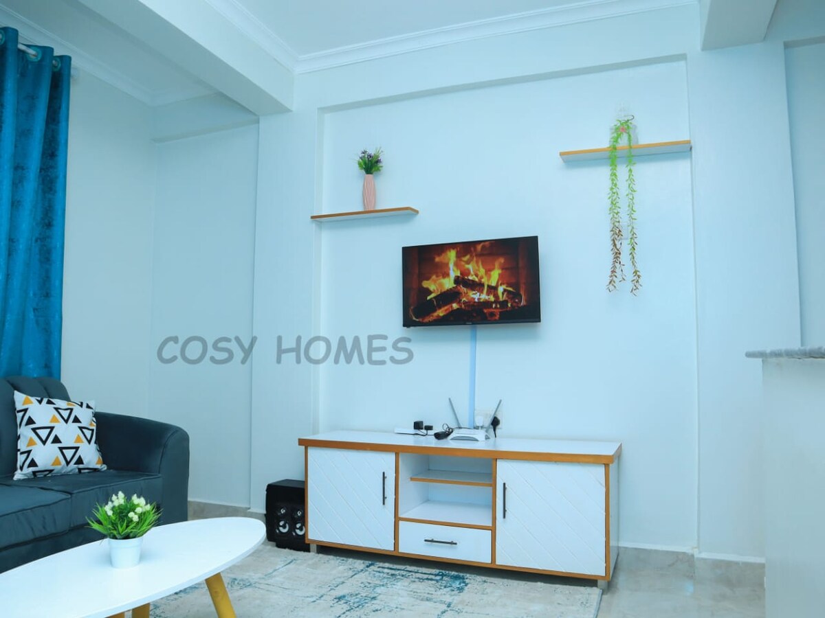 A modern living area features a dark sofa with decorative cushions, complemented by a light wooden entertainment unit. A flat-screen TV is mounted on the wall, displaying a fireplace scene. Shelves with potted plants are displayed above, contributing to a clean, stylish atmosphere.