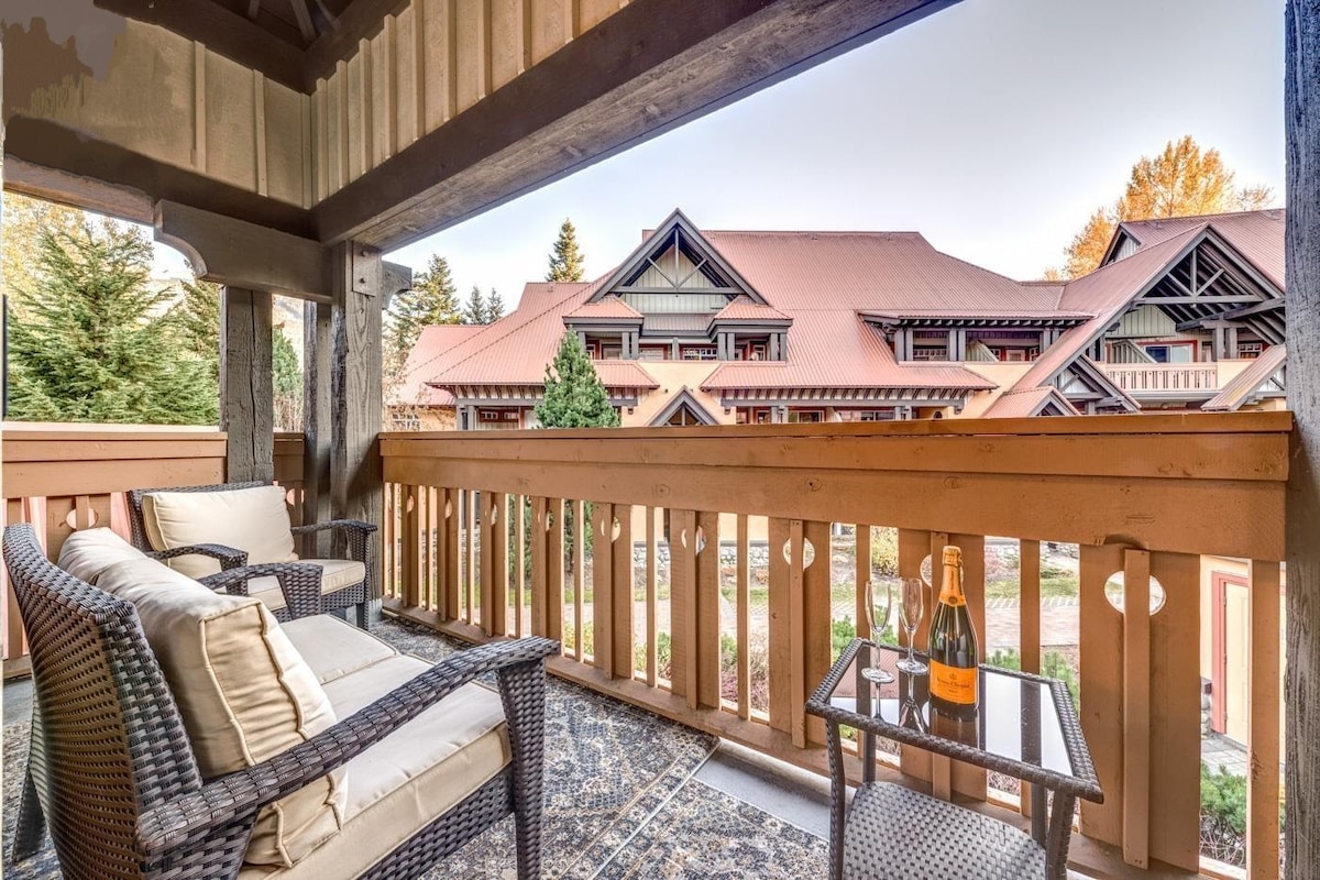 A private balcony is shown with comfortable seating, featuring a small table with a bottle of champagne and glasses. The view includes the surrounding greenery and the elegant architecture of nearby buildings, complemented by a warm ambiance created by natural lighting.