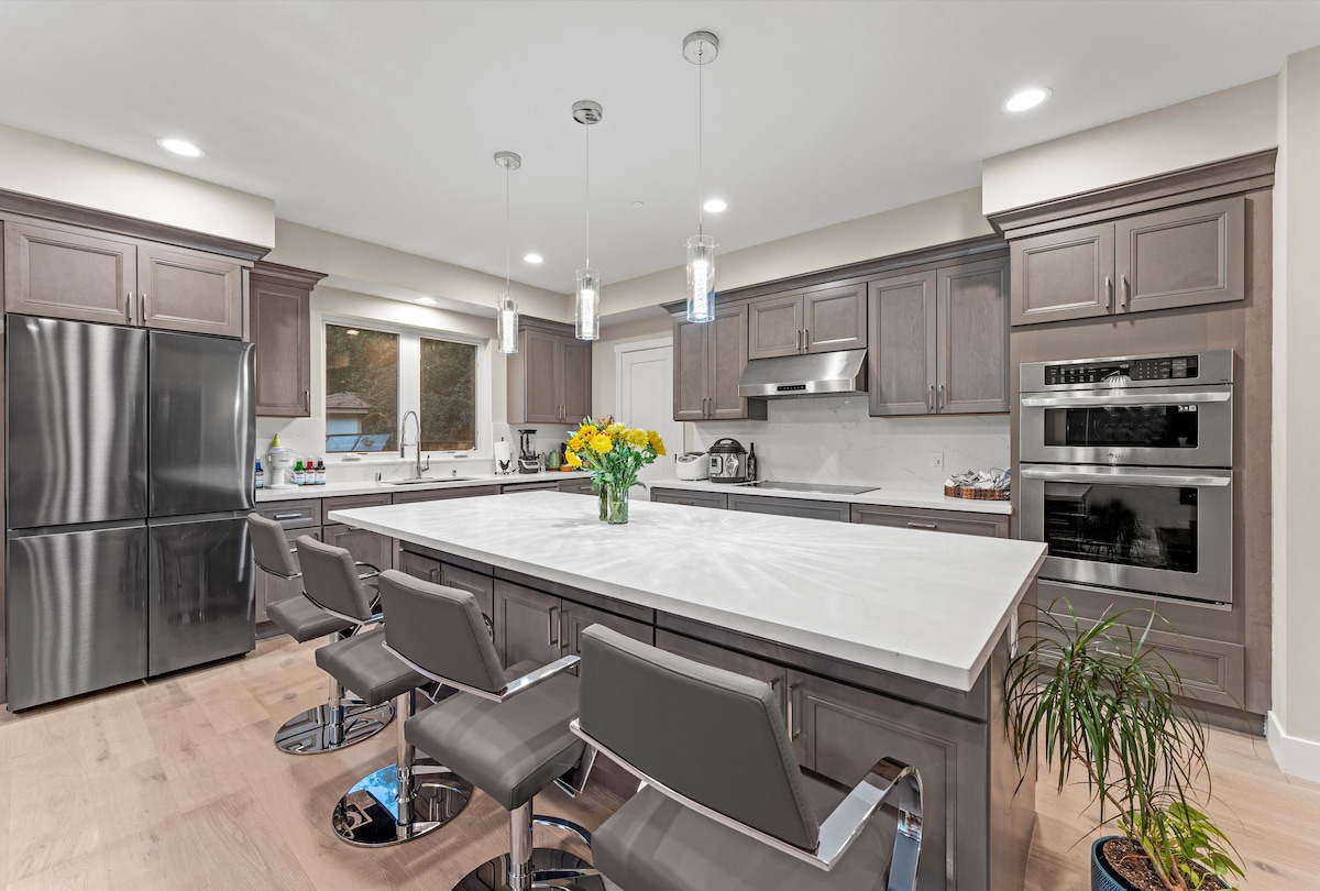 A modern kitchen showcases a large island with polished white countertops, surrounded by sleek gray bar stools. Stainless steel appliances are installed, including a refrigerator and double ovens. Natural light is invited through a window, complemented by a vase of yellow flowers set on the island.