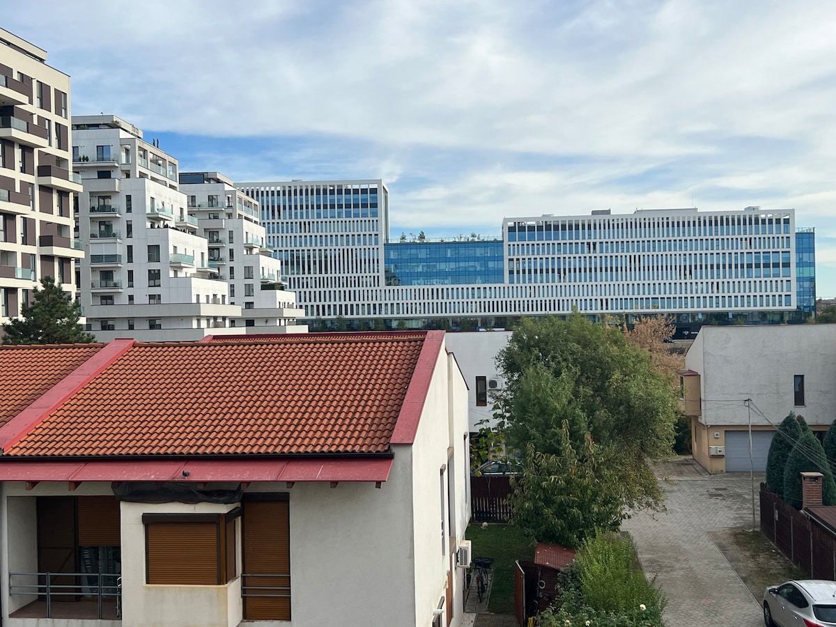 A view from a balcony captures a mix of modern architecture and greenery. The foreground features a building with a red-tiled roof and large windows, while the background showcases a contemporary structure with a glass facade lined with evenly spaced windows.