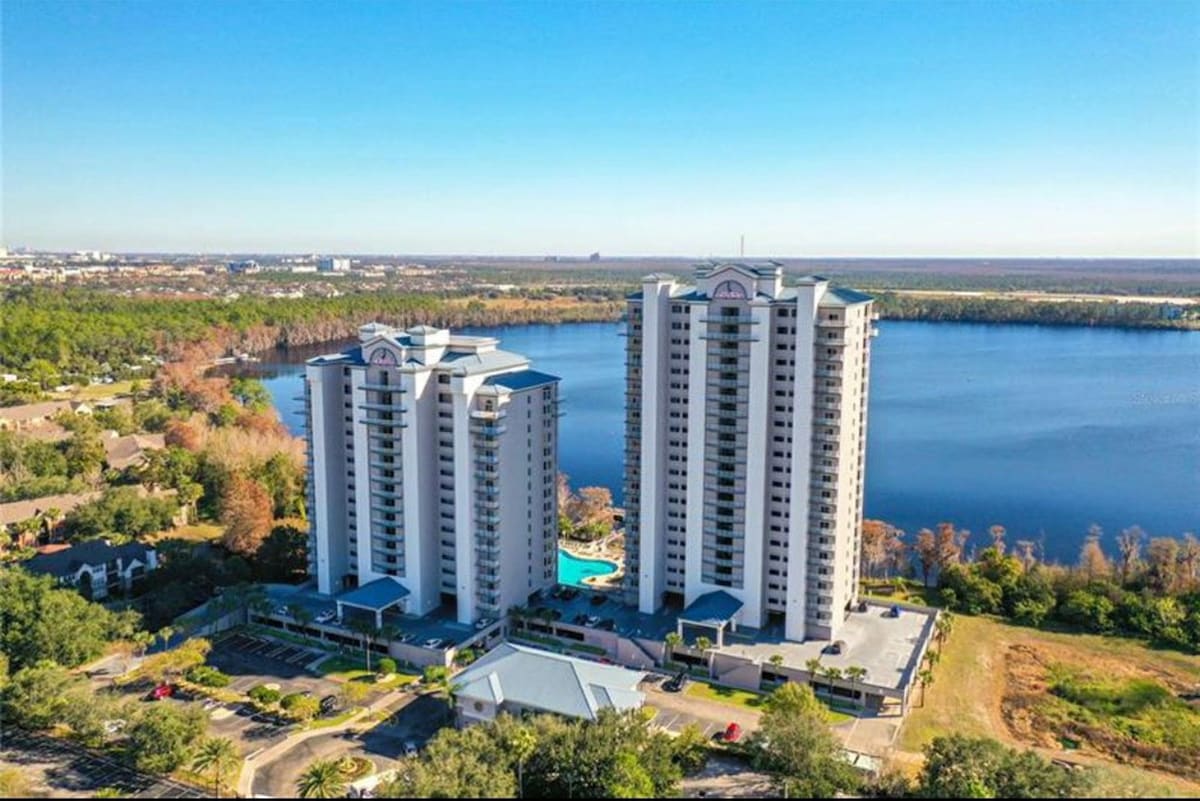 Two tall condominium buildings are situated near the edge of Lake Bryan, showcasing a modern architectural design. A blue pool is visible in the foreground, surrounded by landscaped greenery and parking areas. The expansive lake and distant city skyline create a serene backdrop.