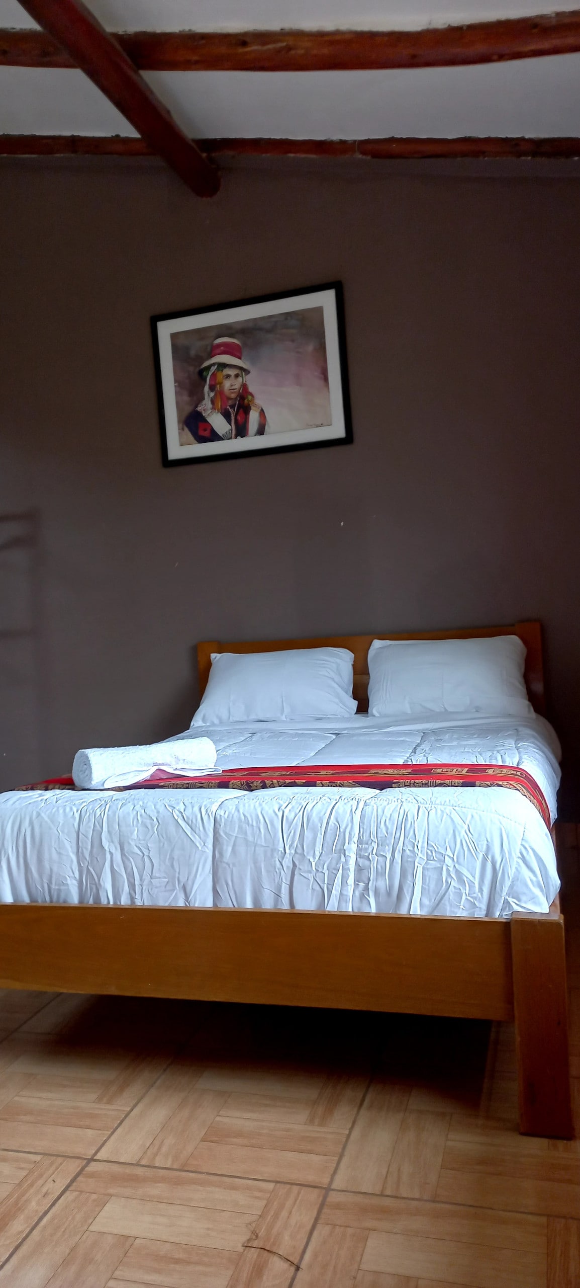 A spacious bed is centered against a textured wall, topped with crisp, white linens and accentuated by a bright red runner. A framed artwork depicting a figure hangs above the bed. Natural light softly illuminates the room, highlighting the wooden beams of the ceiling.