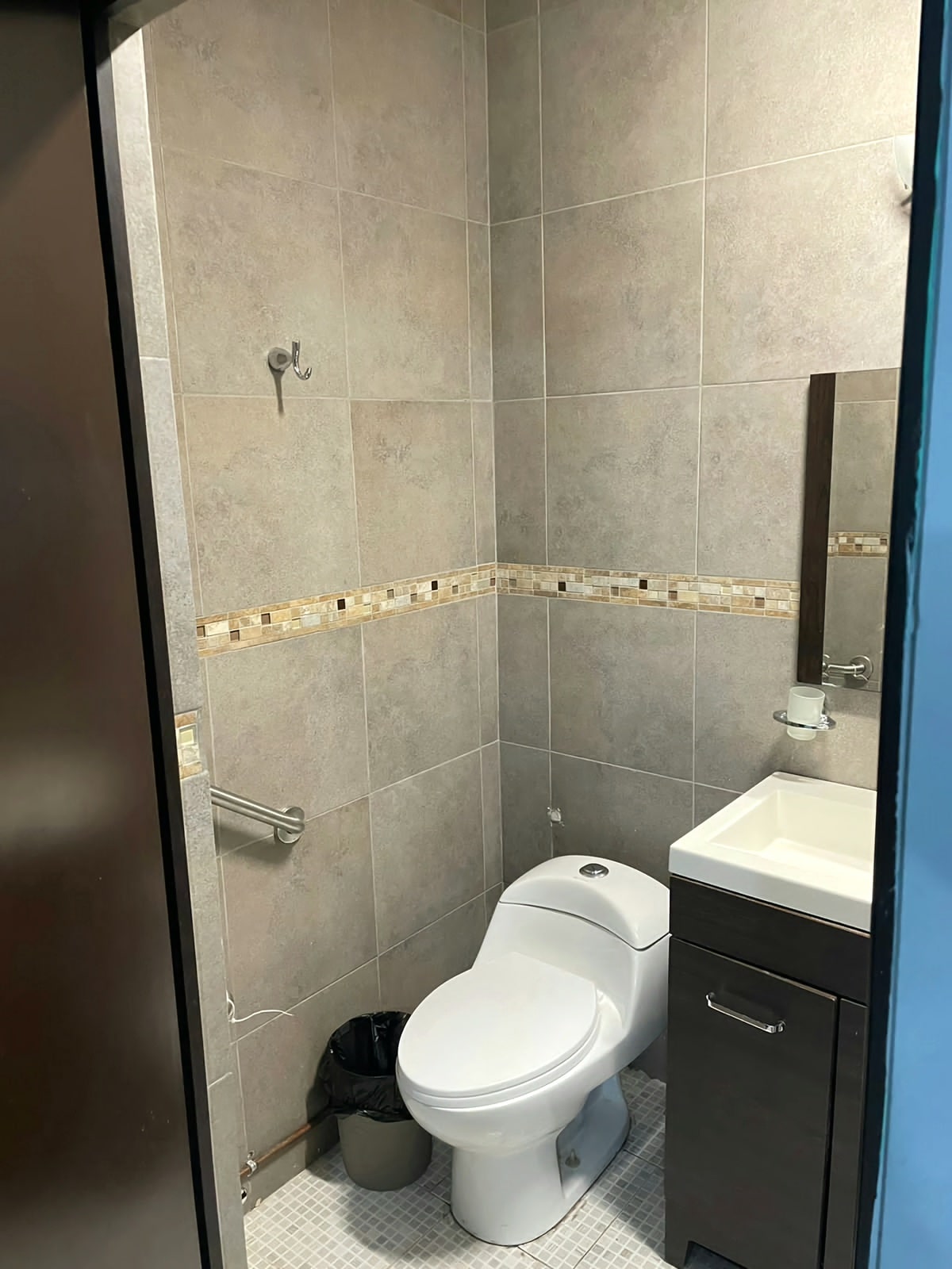 A compact bathroom features gray tile walls and a mosaic accent strip. A toilet is positioned beside a small vanity with a sink and mirror. A trash bin is located on the floor, and a towel hook is mounted on the wall.
