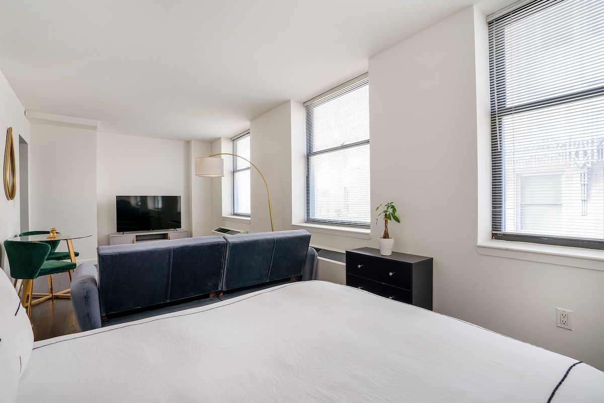 A bright studio room is designed with large windows allowing natural light to flood in. A spacious bed is positioned near a modern sofa and a dark chest of drawers. An elegant floor lamp and a small green chair add a touch of style to the welcoming space.