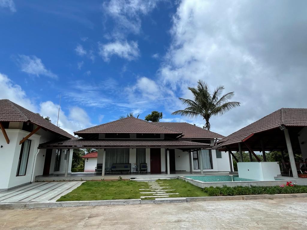 A modern villa is displayed under a partly cloudy sky, featuring a spacious entrance with a stone pathway leading to a well-maintained lawn. The structure includes a patio area with overhanging roofs and a swimming pool adjacent to the house.