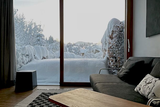 A modern living area features a comfortable sofa positioned near a large window. Outside, a serene winter landscape is visible, with snow-covered trees and a blanket of snow adorning the ground. Soft natural light fills the space, enhancing the cozy atmosphere.