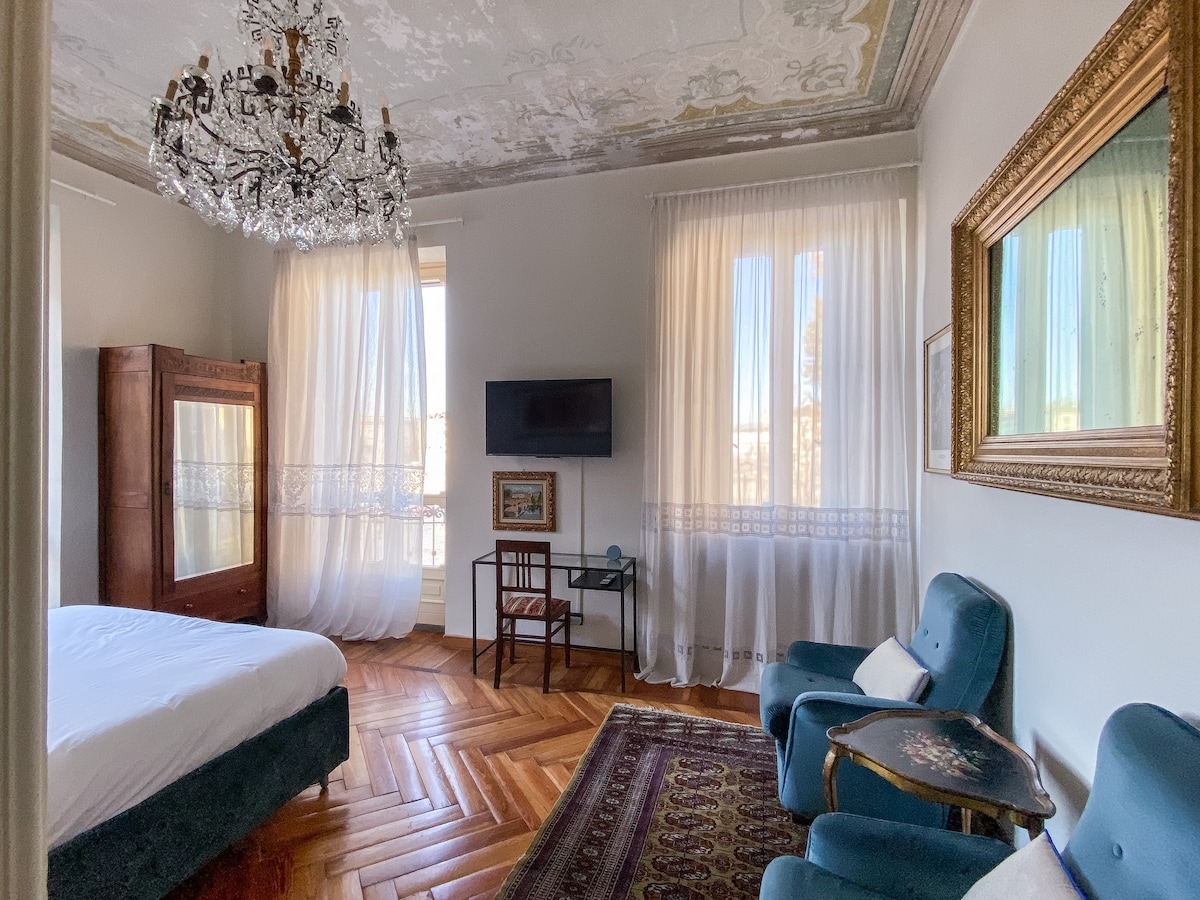 A spacious bedroom features a chandelier and large windows draped with sheer curtains, allowing ample natural light. The interior includes a comfortable bed, vintage furniture, and an elegant mirror, enhancing the room's classic charm. A warm wood floor complements the decor.