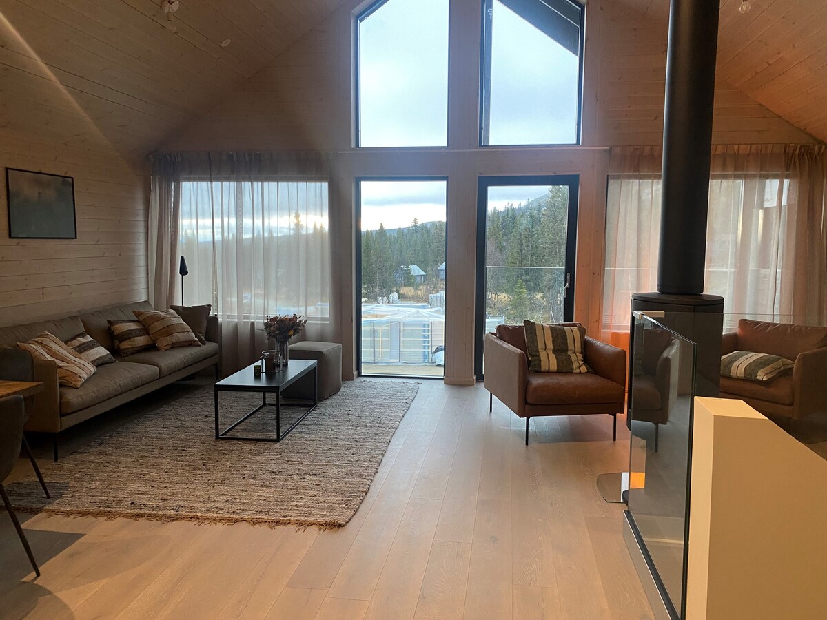A spacious living area is highlighted by large windows allowing natural light to fill the room. A cozy seating arrangement includes a sofa and armchairs, complemented by a coffee table. Light-colored curtains frame the view of the surrounding landscape, enhancing the serene environment.