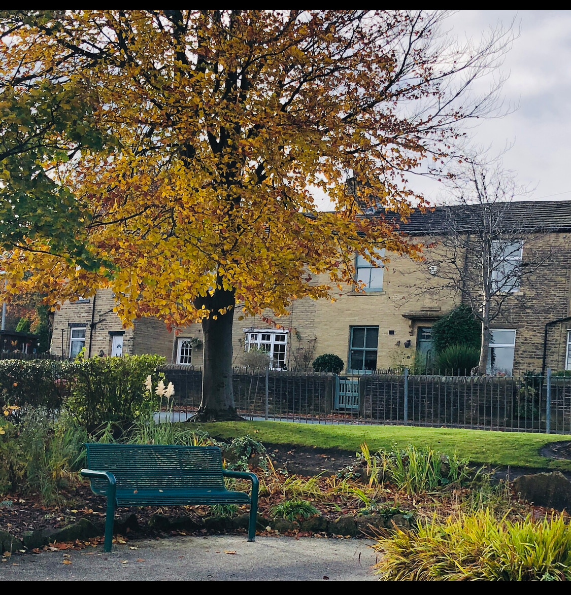 A park scene features a green bench surrounded by autumn foliage, with a large tree displaying golden leaves. In the background, a stone cottage is visible, with multiple windows and a well-kept garden. A low stone wall delineates the property.