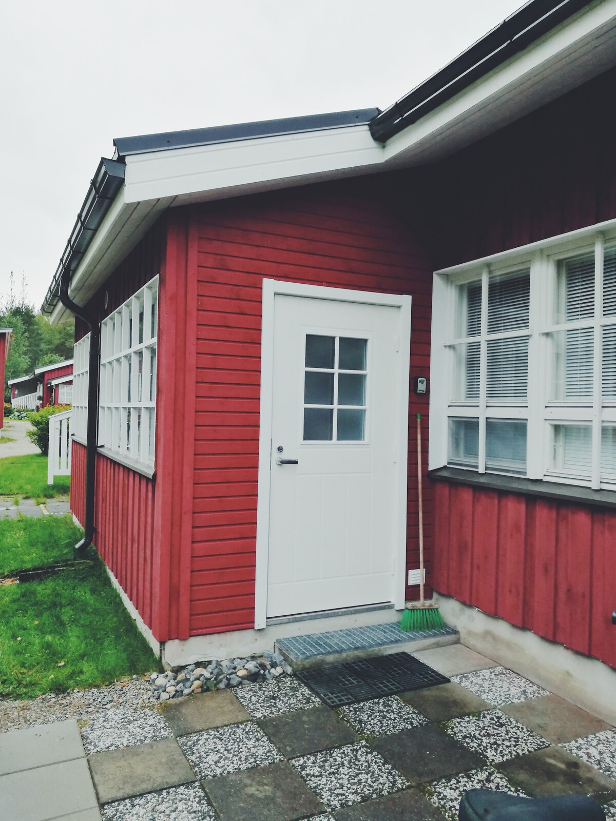 The exterior of a red wooden house features a white door with a grid window design. A clean, paved pathway leads to the entrance, complemented by a green lawn and neatly arranged stones. Large windows provide natural light and a view of the surroundings.