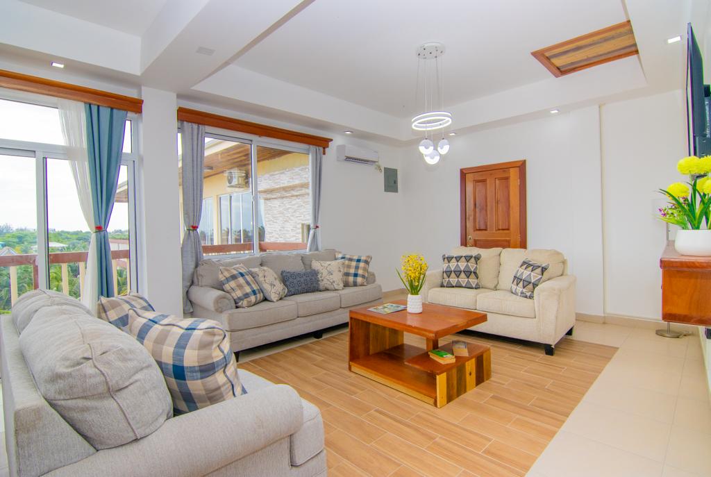 A spacious living area is presented, featuring two light-colored sofas adorned with patterned cushions. A wooden coffee table occupies the center, surrounded by natural light from large windows. Soft blue curtains complement the decor, while greenery adds freshness to the space.
