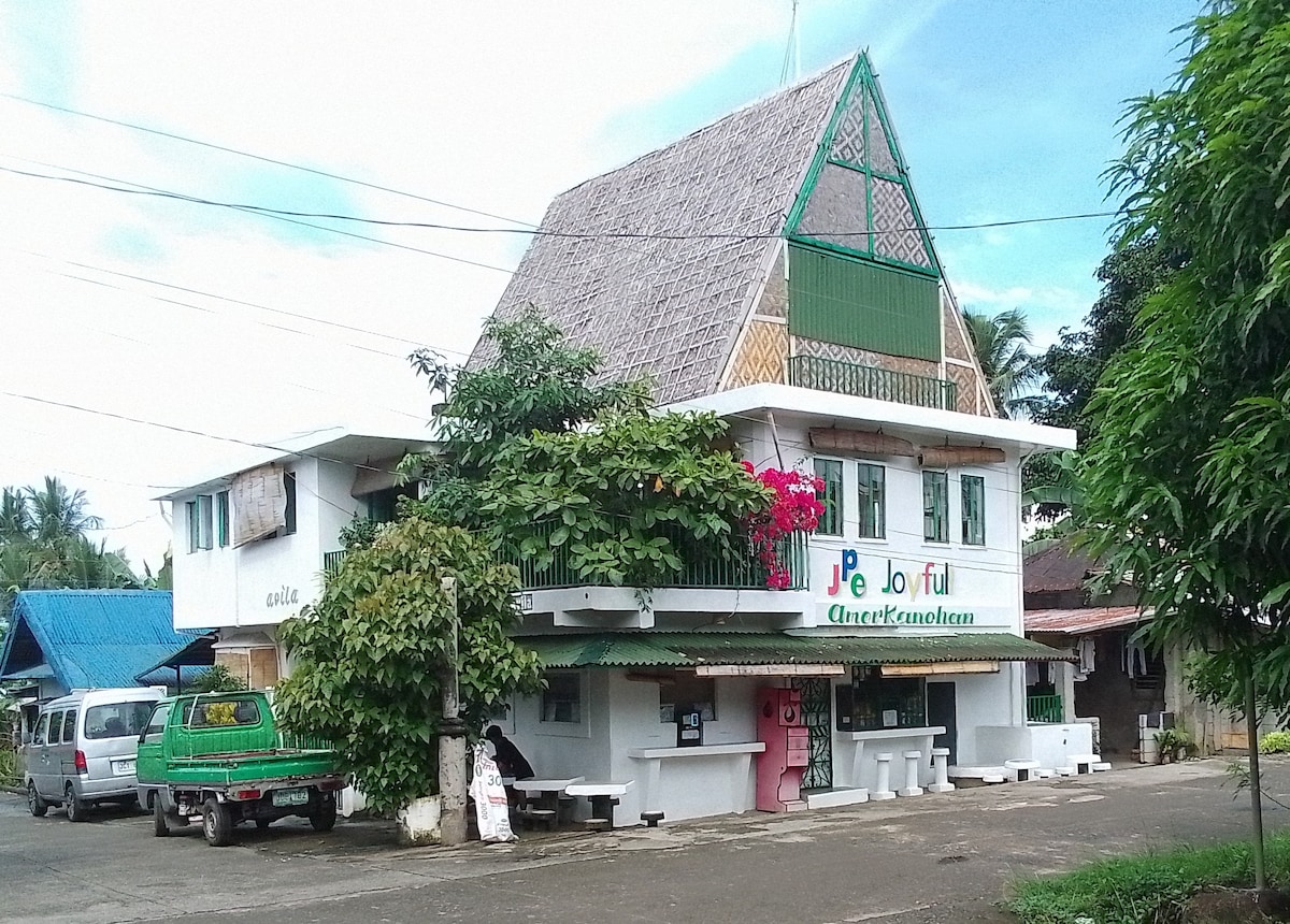 A two-story building with a traditional thatched roof is showcased. Colorful signage is prominently displayed at the entrance, while vibrant greenery frames the facade. The surrounding street features cars parked along the side, indicating a lively neighborhood atmosphere.