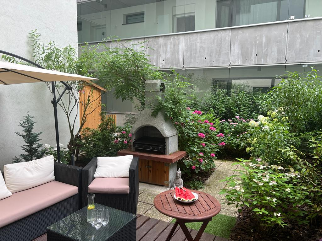 An outdoor terrace features comfortable lounge seating and a barbecue area framed by lush greenery. A round table is accompanied by two glasses and a small plate with fruits. A serene atmosphere is created by flowering plants and a stone fireplace.