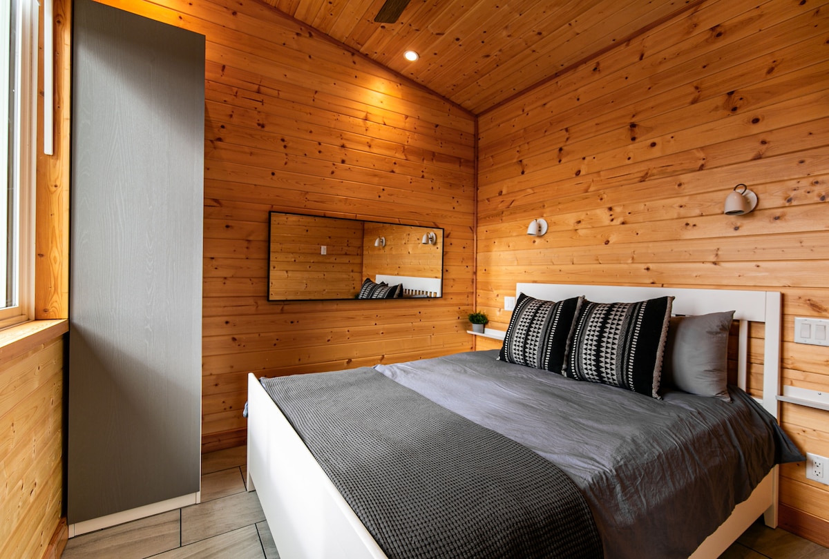 A cozy sleeping area features warm wood-panelled walls and a spacious bed dressed in gray linens. Two decorative pillows complement the bedding. Natural light streams in from a large window next to a sleek wardrobe, with a mirror reflecting the inviting interior.
