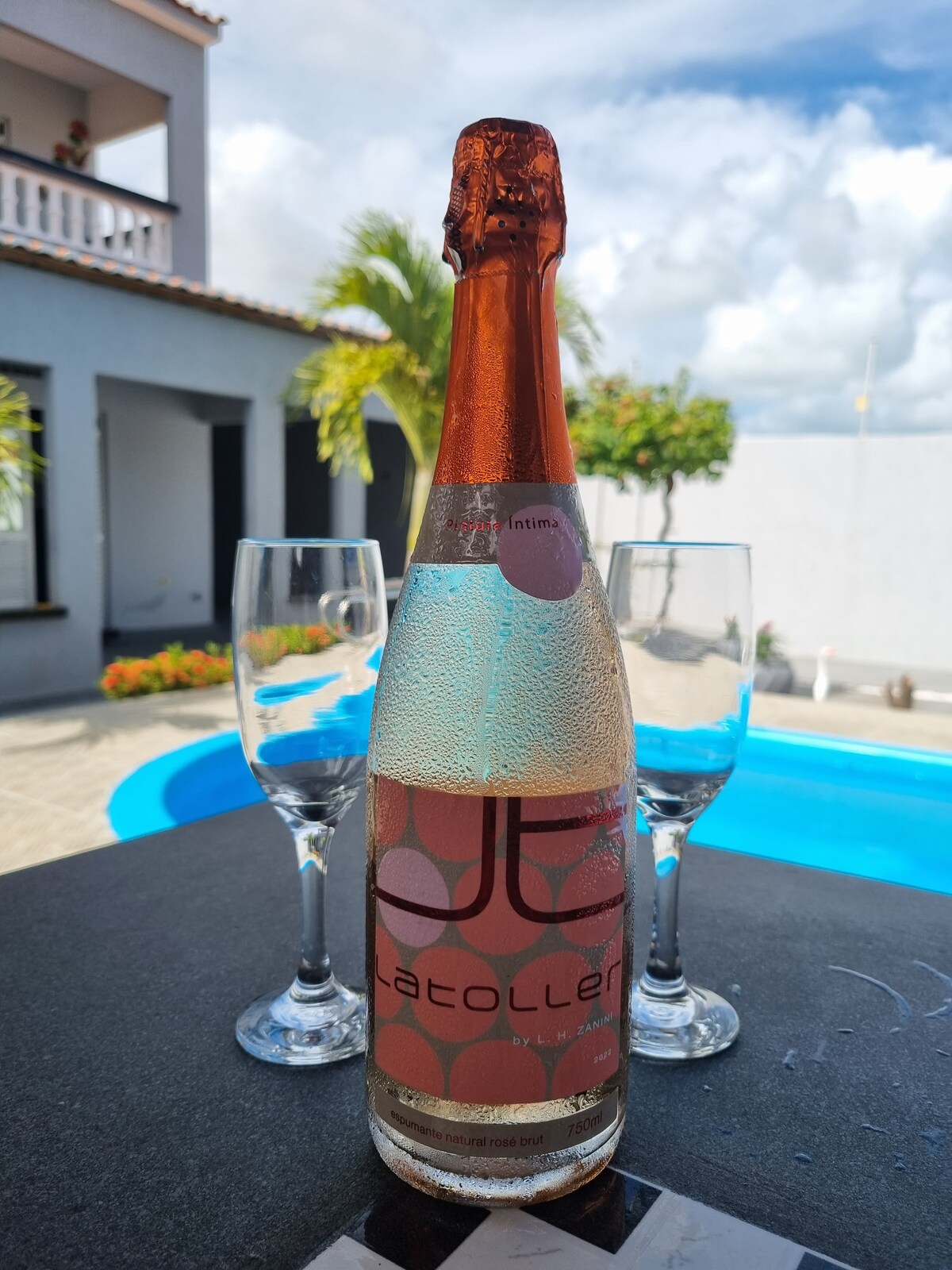 A chilled bottle of sparkling wine is displayed on a table next to two clear glasses, with droplets of condensation visible on the bottle. In the background, a vibrant blue pool is partially visible, surrounded by lush greenery and a bright sky.