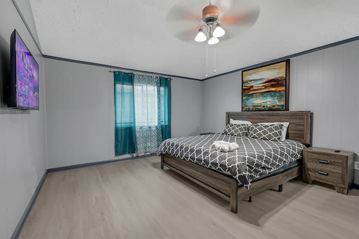 Ეს არის თანამედროვე საძინებელი, რომელშიც დგას საწოლი, რომელშიც დგას შაბლონური პლედი, ხის ტუმბო და კომოდი, ლამინირებული იატაკი და კედელზე დამაგრებული ბრტყელეკრანიანი ტელევიზორი.