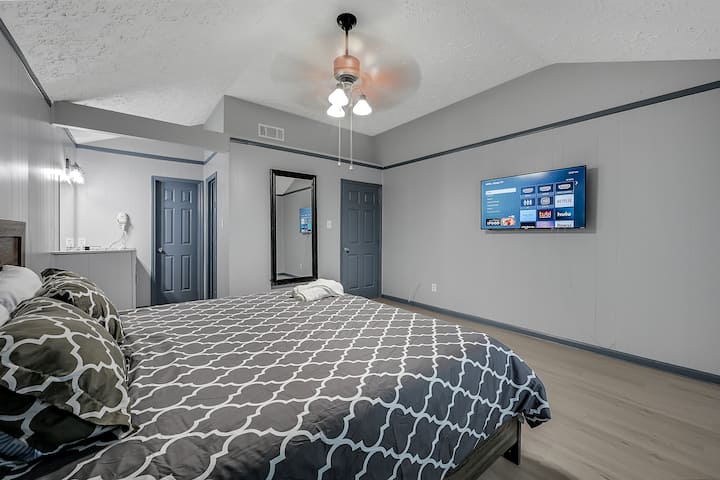 Თანამედროვე საძინებელი, რომელშიც დგას დიდი საწოლი შაბლონური პლედით, თანამედროვე ნაცრისფერი კედლები თეთრი მორთვით, ხის იატაკი და ბრტყელეკრანიანი ტელევიზორი. Მოყვება ფლიგელის სააბაზანო და სარკისებური კარადა.