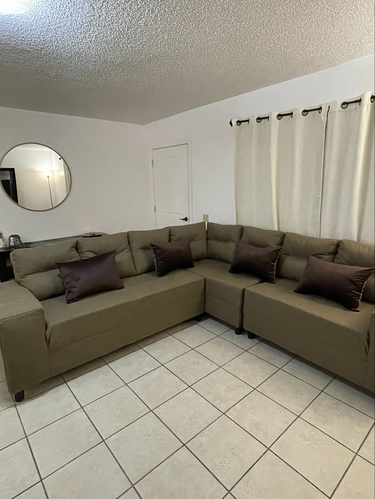 A spacious corner couch in a neutral tone features several throw pillows in rich brown hues. Light reflects softly off a round wall mirror, while light beige curtains frame a nearby window. The tiled floor adds to the clean and simple aesthetic of the living space.