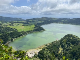 A panoramic view captures a vast lake surrounded by lush green hills and valleys. The water reflects varying shades of green, with hints of blue under the sky. The distant landscape reveals rolling hills and scattered trees, creating a serene natural environment.